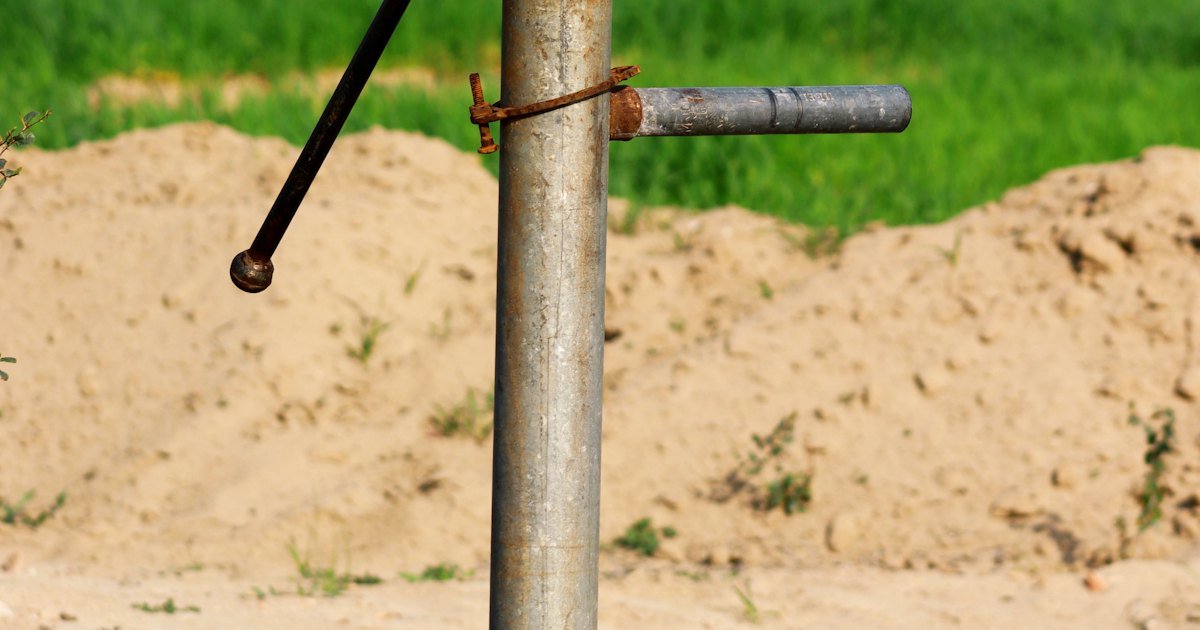 A metal groundwater hand pump pipe in front of a sand heap and green field